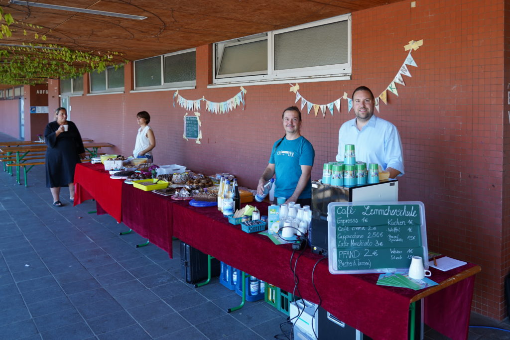 Verkaufsstand „Café Lemmchenschule“ mit Kuchenbuffet, Kaffeemaschine und Preistafel unter dem Vordach