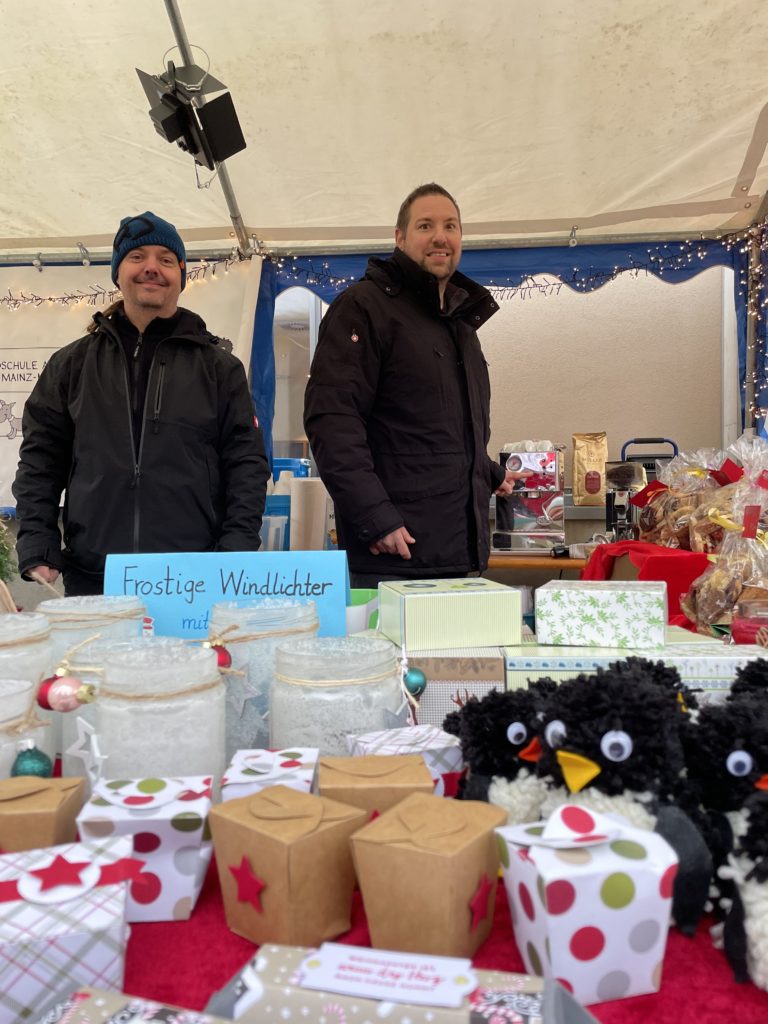 Zwei Ehrenamtliche hinter Windlichtern, Bommel-Pinguinen und Geschenkschachteln, Schild „Frostige Windlichter“